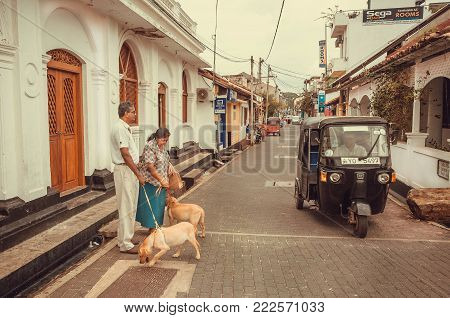 Galle, Sri Lanka - Dec 23, 2017: Elderly Couple With Dogs Walking Past Tuk-tuk Driving In Historical