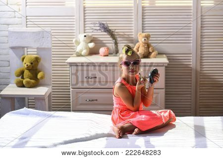 Makeup and kids fashion concept. Little girl paints lips with lipstick sitting in childroom. Child wears nice dress and sunglasses and takes cosmetics. Girl with curlers smiles in room with toys.
