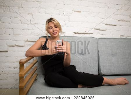Smiling hipster girl chatting in social network via mobile phone while sitting on a comfortable sofa. Happy woman reading text message on portable cell telephone, sitting on couch in modern interior