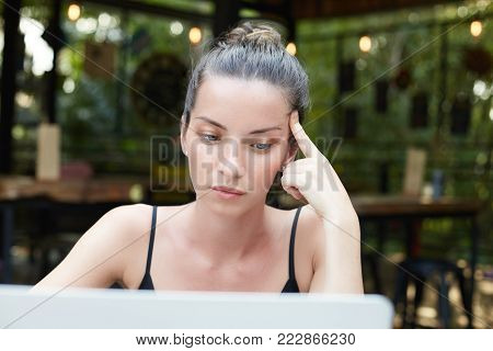 Portrait of beautiful mixed race brunette woman with well-shaped eyebrows, green shining eyes and full lips touching temple thoughtfully, having hair in bun sitting at modern cafe working with laptop.