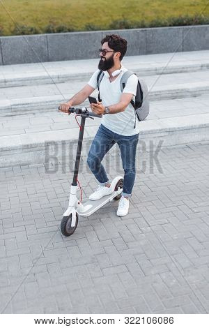Bearded Man With Smartphone Stepping On Modern Electric Scooter And Looking Away While Standing On P