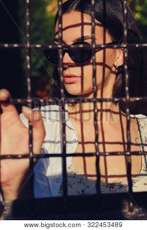 Portrait Of Girl In Sunglasses Posing In City Behind A Trellised Fence. Dressed In Top With Floral P