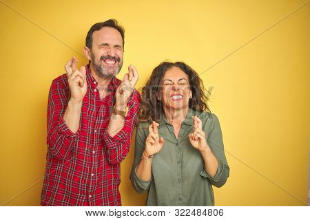 Beautiful middle age couple over isolated yellow background gesturing finger crossed smiling with hope and eyes closed. Luck and superstitious concept.