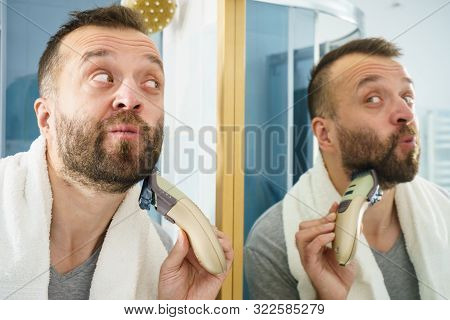 Bearded Man Looking At Himself In Mirror Trimmng, Shaving His Beard Using Electric Timmer Razor.