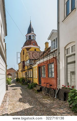 A Bike And Hollyhocks In A Narrow Cobblestone Alley And A Bell Tower In The Background, Faaborg, Den