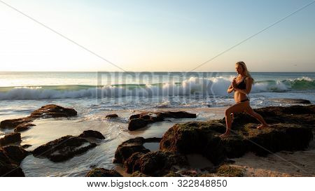 Young Woman Practicing Yoga, Standing In Virabhadrasana Pose On The Beach. Sunset Time. Yoga Retreat