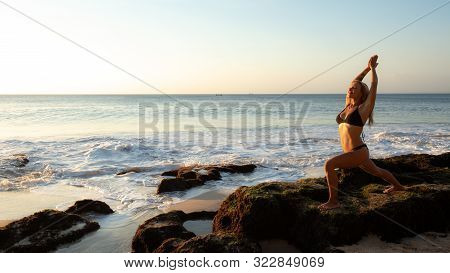 Young Woman Practicing Yoga, Standing In Virabhadrasana Pose On The Beach. Sunset Time. Yoga Retreat