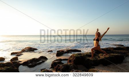 Young Woman Practicing Yoga, Standing In Virabhadrasana Pose On The Beach. Sunset Time. Yoga Retreat