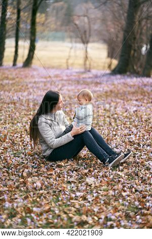 Mom Sits On Fallen Leaves In The Park, Holding The Baby On Her Lap, Sitting With Him Face To Face