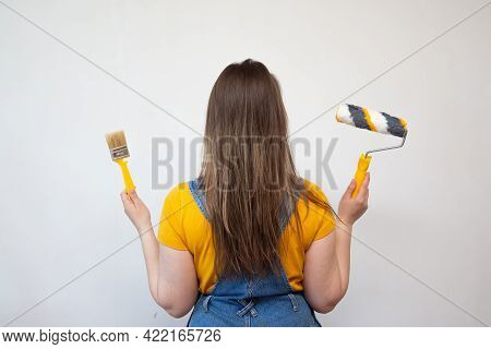 Unsure Woman Chooses Between Roller And Brush On Background Ladder And Gray Cement Wall. Choice Of T