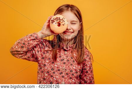Little girl playing and holding donut as glasses close to eyes isolated on yellow background with copyspace. Pretty female kid with sweets