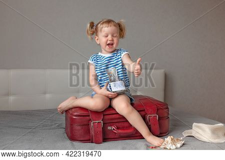 Small Happy Laughing Blonde Girl In Striped Blue And White T-shirt And Blue Shorts Sits Burgundy Sui