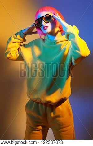Modern girl with bright pink hair poses in yellow sunglasses and yellow suit in mixed color light. Sport chic fashion. Youth fashion and beauty trends. 