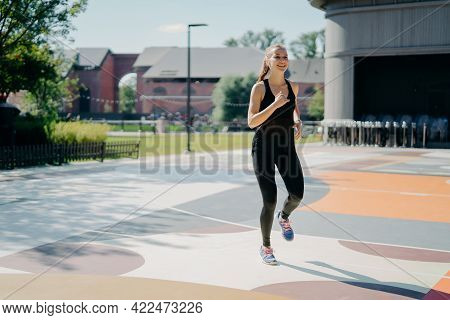 Cheerful Athletic Woman Runs Actively On Stadium Dressed In Black Sportclothes Enjoys Physical Activ