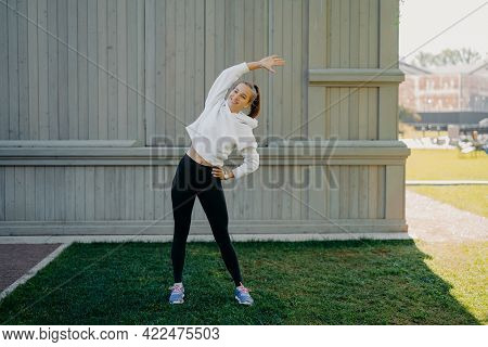 Full Length Shot Of Active Sportswoman Does Exercises Outdoors Stetches Body Leans At Different Side