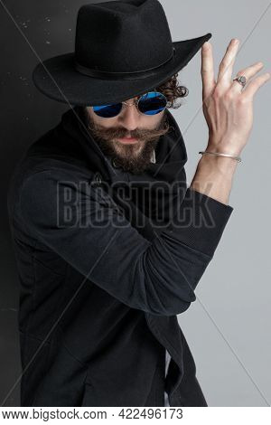mysterious latin fashion model with long beard and moustache pushing hat up and confidently posing while looking down in studio