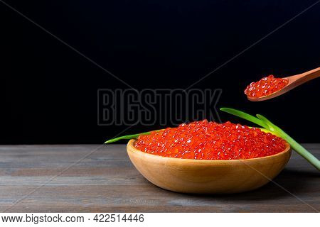 Red Caviar In A Wooden Cup On A Black Background With A Spoon And Green Onions. A Large Pile Of Brig
