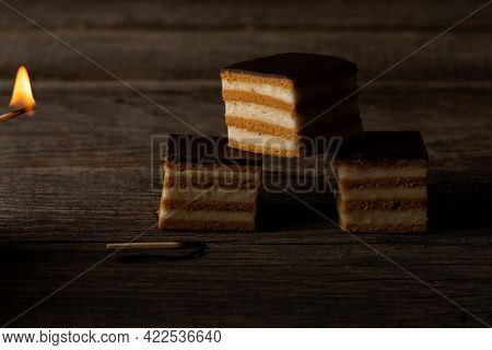 Three Slices Of Gerbeaud Cake, Lit By The Fire Of A Burning Match, Are Lying On Weathered Wood Plank