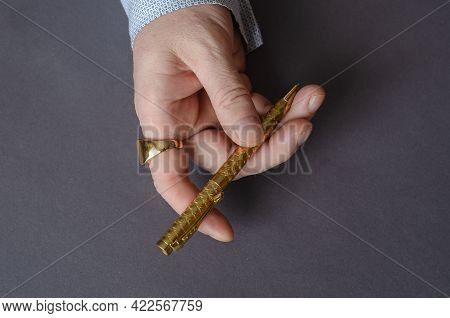 A Man With A Gold Ring Is Holding A Yellow Metal Pen. The Right Hand Of An Adult Male With A Signet 