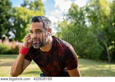 Portrait of mixed race sports man resting after run at park with copy space while listening to music. Middle adult indian jogger adjusting wireless earphone while running outdoor. Active mature man.