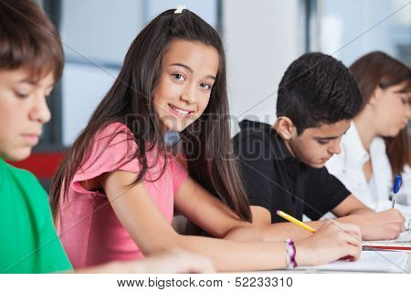 Portrait of happy teenage girl sitting with classmates studying at desk in classroom