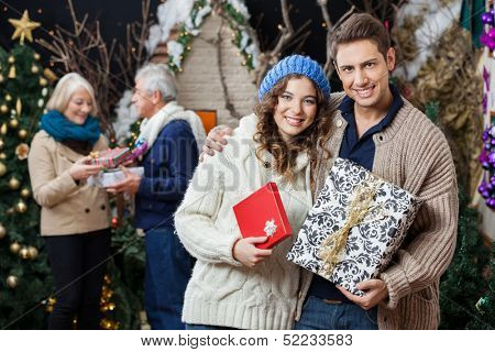 Portrait of happy young couple holding Christmas presents with parents standing in background at store