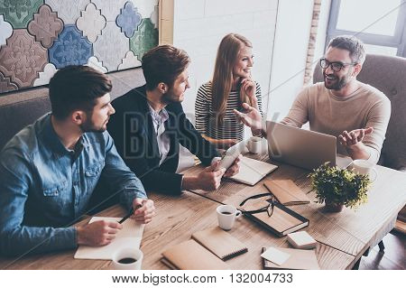 So what we should do next? Top view of young handsome man in glasses gesturing and discussing something with his coworkers with smile while sitting at the office table