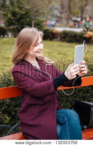 Happy charming young woman listening to music and making selfie using tablet in park