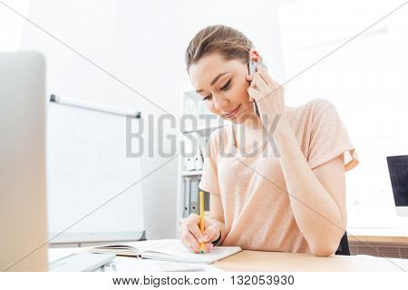 Smiling cute young woman talking on mobile phone and writing in office