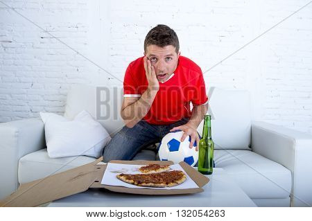 young man alone holding ball with beer and pizza in stress wearing team jersey watching football game on television at home living room sofa couch excited in disbelief face expression