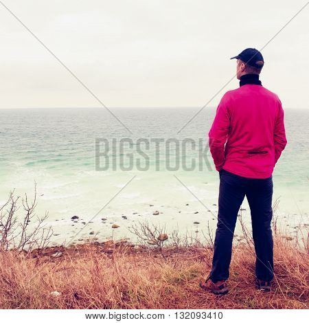 Man In Windcheater Stand On Cliff Above Bay And Looki At Sea Far Horizon. Windy Cold Weather