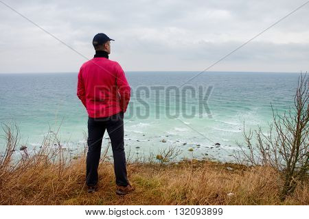 Man In Windcheater Stand On Cliff Above Bay And Looki At Sea Far Horizon. Windy Cold Weather