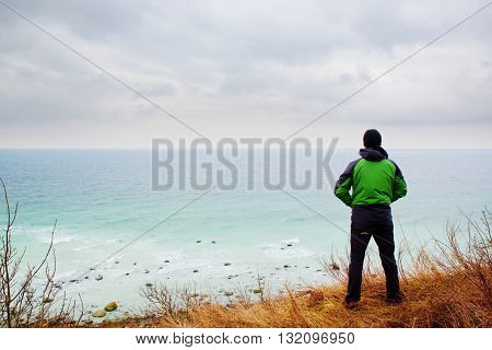 Man In Green Windcheater Stand On Cliff Above Bay And Looki At Sea Far Horizon.