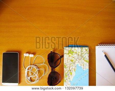 travel concept flay lay, sunglasses, map, headphones, phone, pencil, copybook, table, wooden surface