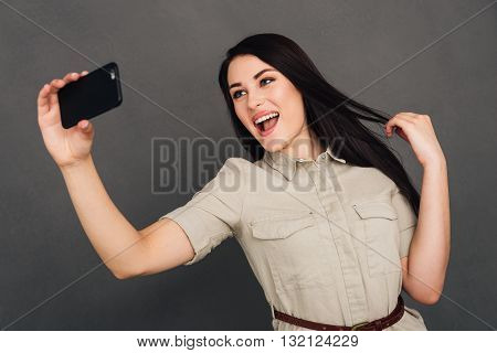 Capturing fun. Beautiful young woman making selfie by smart phone and smiling while standing against grey background