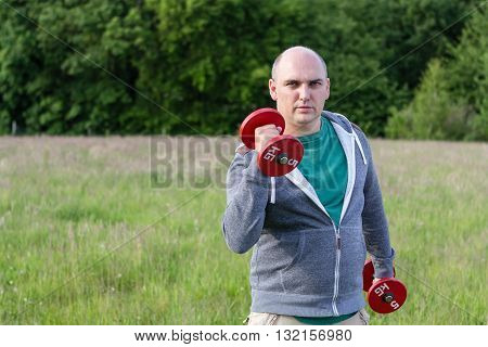 Man working outdoors with 5kg dumbbells. He holds one up.