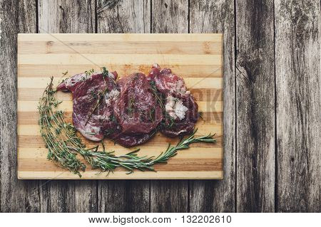 Fresh beef steak ready for BBQ cooking. Raw meat on a cutting board with rosemary leaf top view at rustic wood. Raw beef, veal meat on wood, closeup. Marinated in spices raw steak for barbecue.