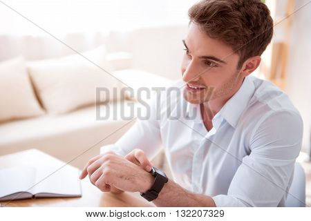 Positivity in mind. Pleasant cheerful handsome man sitting at the  table and using his smart watch while sitting at the table