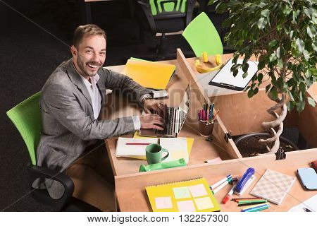 Portrait of happy businessman working on laptop compuer in office. Happy man in grey business suit looking at camera and smiling.