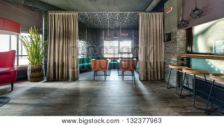 Room in a loft style in a mexican restaurant. On the left there is a crimson armchair with a glass table near it and a red lampshade over it. Big cactus in a wicker pot is behind the armchair. On the right there is a wooden rack with wooden chairs 