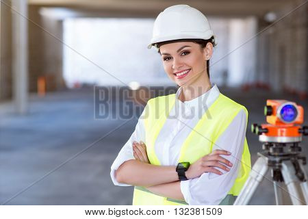 Modernizing. Smiling and content young architect standing and looking at a camera with a geodetic level in a background