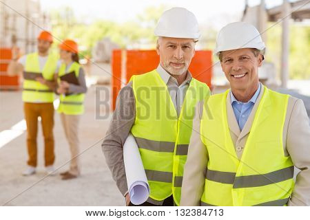 Happy to cooperate together. Content and positive pair of architects standing together and looking at a camera while holding a construction plan with a pair of another architects in a background