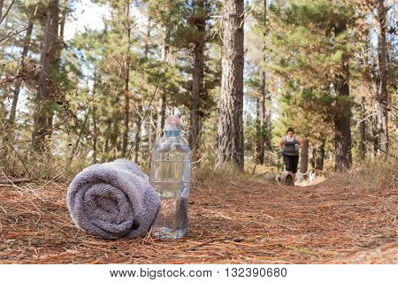 Overweight woman jogging on forest trail with waterbottle and towel in the foreground