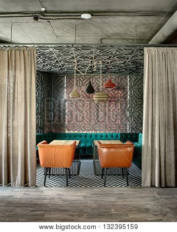 Wonderful interior in a loft style in a mexican restaurant. There is a zone decorated with metal lattices and curtains. In the zone there are green sofas, brown chairs, wooden tables and multi-colored lamps over them. On the floor there is a gray parquet