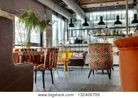 Charming hall in a loft style in a mexican restaurant with open kitchen on the background. In front of the kitchen there are wooden tables with multi-colored chairs and sofas. On the sofas there are color pillows. In the kitchen there is a rack