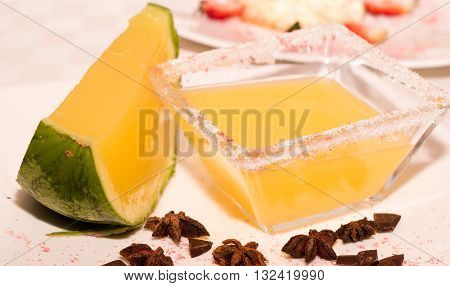 Square transparent bowl with sugarcoating around edges and yellow liquid inside nexto to melon.