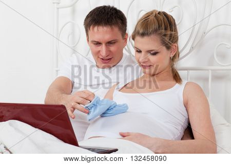 Happy pregnant family with notebook and children's bootees lies on the house bed.