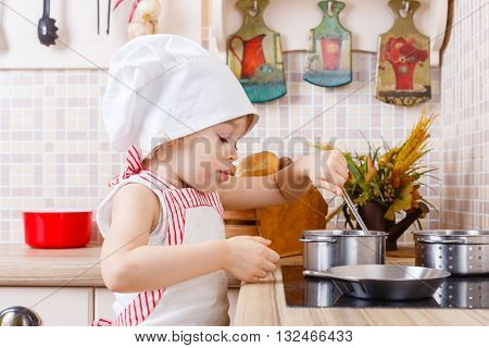 Little girl in apron and cap of the cook stands in the kitchen near cooker in the house. Mother's helper. 2 year old.