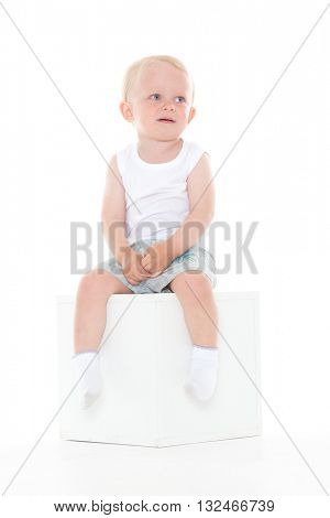Unhappy yearling baby boy sits on cube on a white background.