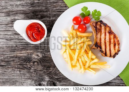 grilled pork chops on a white dish with french fries cherry tomatoes cilantro leaves and ketchup on a wooden table blank space left top view